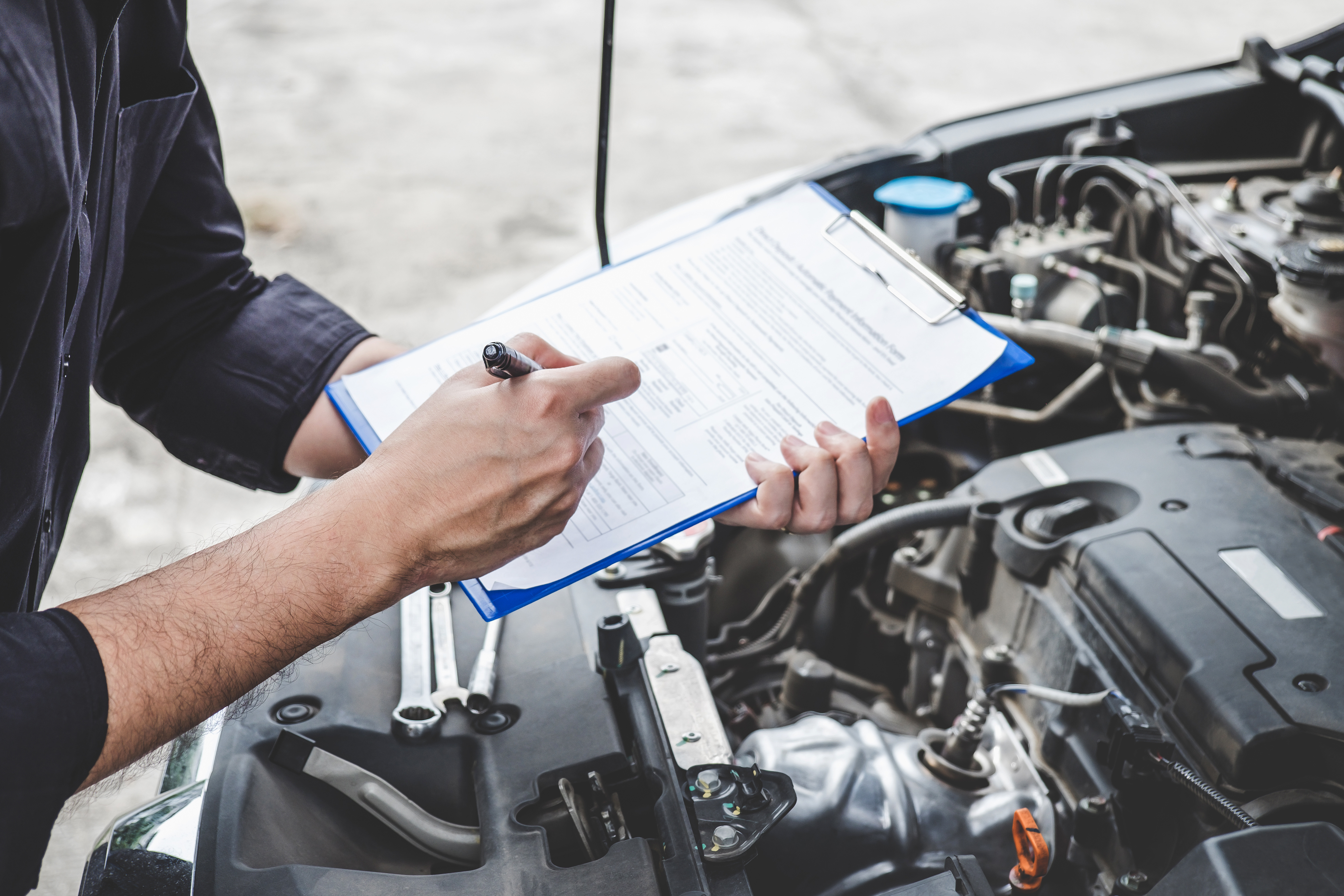A mechanic holds a clipboard over the open bonnet of a car and fills out a checklist.
