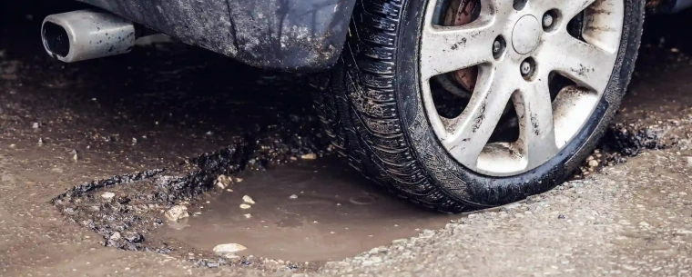 A dirty car with its back tyre in a deep pothole with a puddle in it.
