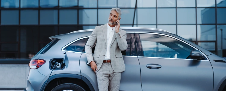 A businessman in a grey suit takes a call while waiting for for his new plug-in hybrid car to charge.