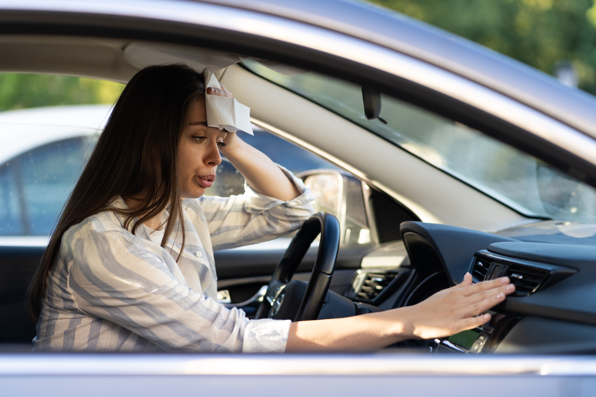 A woman holds a paper towel up to her head and tries to make her air conditioning work in a hot car.